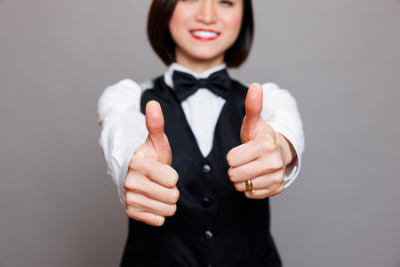 Cheerful waitress showing thumbs up gesture with closeup on arms. Young asian woman receptionist sharing restaurant service positive experience with approval symbol in studioの写真素材