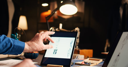 Old couturier checking fashion sketches in atelier, working on modern designs with laptop and tablet. Experienced designer searching handmade models online, needlework. Handheld shot.の写真素材