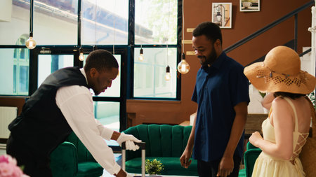 Male tourist giving tip to bellboy helping with luggage, paying cash for luxury front desk services. Young guest thanking bellhop for carrying trolley bags, money for assistance.の写真素材