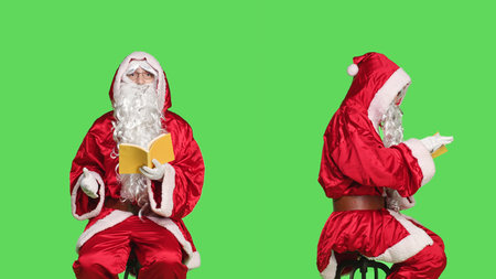 Saint nick reads lecutre novel on chair, man dressed as santa claus with red suit and glasses while he is reading literature book over greenscreen. Person in costume enjoying fairytale fiction.の写真素材