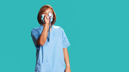 Asian experienced nurse doing mock up showing of properly using professional stethoscope. Knowledgeable clinic worker demonstrating medical gear usage, isolated over blue studio backgroundの写真素材
