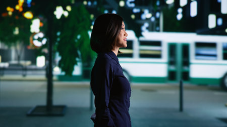 Asian woman strolling around city on sidewalk, enjoying urban promenade under streetlamps. Confident person walking at nighttime, admiring town landscapes and skyscrapers. Handheld shot.の写真素材