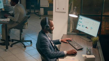 Company executive enjoying music while working on business revenue statistical charts in modern office. Relaxed accountant with headphones on researching key data for company project during nightshiftの写真素材