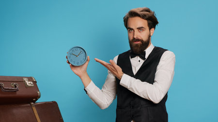 Doorkeeper shows wall clock on camera, checking time and trying to be punctual in hotel travel sector. Man hotel concierge using watch to see hour and minutes in studio next to trolley bags.の写真素材