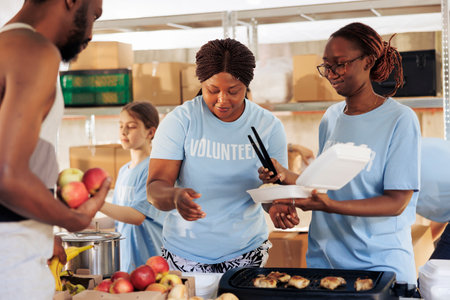 Friendly black women happily serving free food to the poor and needy african american man at a homeless shelter. Hungry, less fortunate male individual receives meal donation from hunger relief team.の写真素材