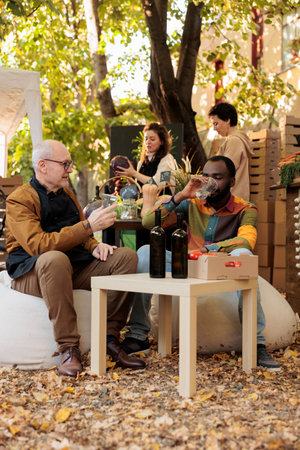 Cheerful people clinking glasses of organic wine at local street festival, tasting homemade wine from bottle at farmers market stand. African american man and elderly person drinking bio product.の写真素材