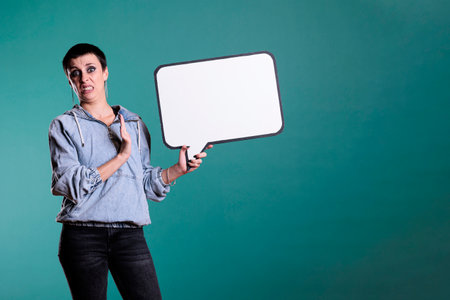 Cheerful brunette woman pointing at empty white speech bubble during studio shot, advertising copy space accessory for text message. Smiling model looking at camera while showing blank template boardの写真素材