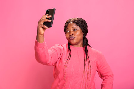 Portrait of lovely woman with stylish hairstyle taking selfie using smartphone while doing funny expression in studio over isolated background. Cheerful attractive person enjoying making photosの写真素材