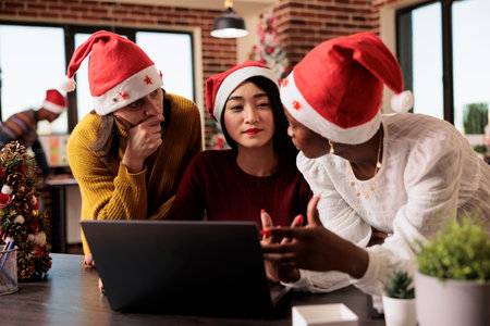 Women workers team gathered around laptop, discussing project tasks in office during christmas holiday. Diverse colleagues in santa hats collaborating on annual report on xmas eveの写真素材
