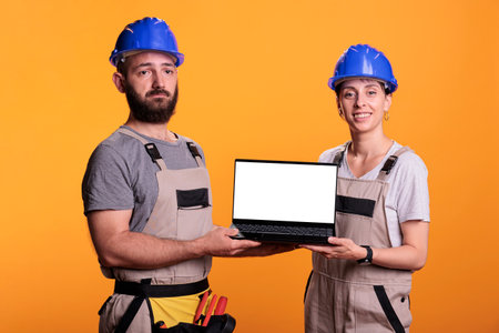 Team of builders showing white empty screen on laptop, wearing overalls and hardhats in studio. Construction workers advertising isolated copyspace display on portable computer, blank mockup.の写真素材