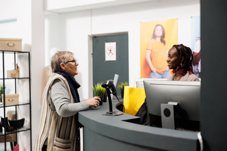 Elderly customer discussing with clothing store employee at cash register, paying for fashionable clothes in shopping centre. Shopaholic woman buying stylish merchandise in modern boutiqueの写真素材