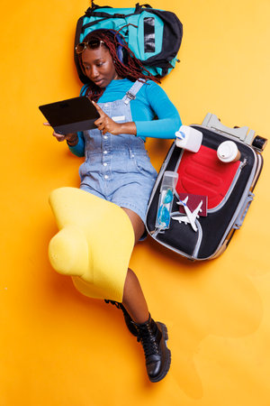 Young person watching film laying down with travelling essentials and trolley bags, preparing to leave on holiday trip. Female tourist sitting on the floor and looking at movie on tablet.の写真素材