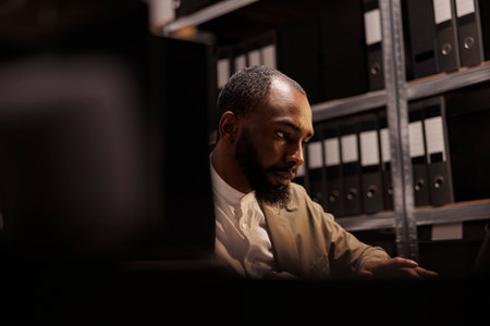 African american cop conducting investigation in police archive, sitting near shelf full of folder...