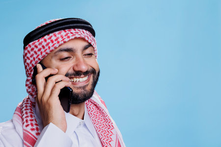 Smiling muslim man wearing ghutra headscarf with rope band talking on mobile phone. Cheerful arab...