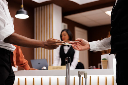Guest giving cash to bellboy in lobby, paying money for luxury service to carry luggage and suitcases. Hotel concierge receiving payment from tourist, helping with trolley bags. Close up.の写真素材