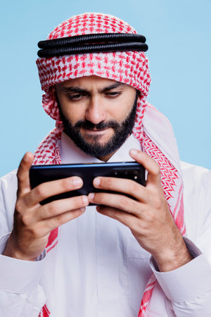 Concentrated muslim man wearing ghutra headscarf playing video game on smartphone. Focused arab gamer dressed in traditional clothes enjoying videogame application on mobile phoneの写真素材