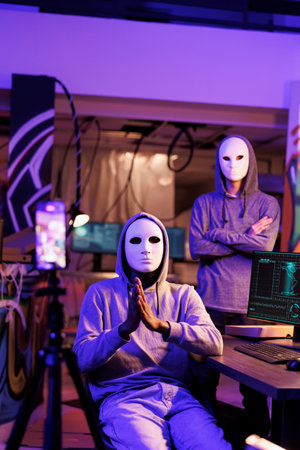 Hackers in masks recording threatening message and demanding victim for confidential privacy information. Anonymous internet scammers doing criminal activities and streaming on mobile phone cameraの写真素材