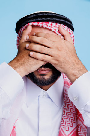 Man dressed in islamic clothes closing eyes with hands while showing see no evil three wise monkeys sign. Young muslim person showcasing eyesight problem concept closeup in studioの写真素材