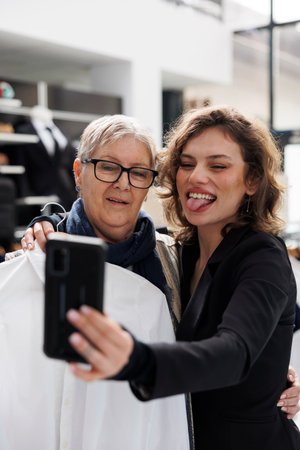 Women taking photo with phone in clothing centre, senior woman holding white shirt in front of camera. Shopaholic clients having fun in modern boutique while buying stylish clothesの写真素材