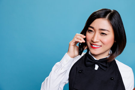 Hotel smiling cheerful asian woman receptionist wearing uniform speaking on smartphone. Joyful waitress answering call and chatting with catering service customer on mobile phoneの写真素材