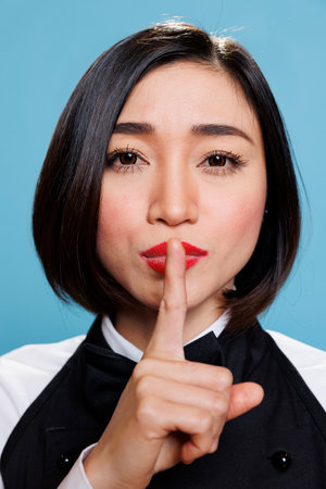 Serious asian woman in waitress uniform holding forefinger on lips while showing tsss gesture and looking at camera. Young receptionist asking for silence closeup portraitの写真素材