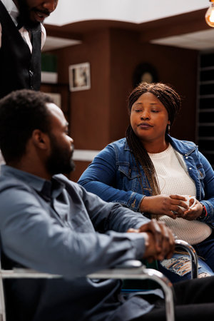 Exhausted young African American couple sitting at hotel lobby ordering coffee, talking with waiter....