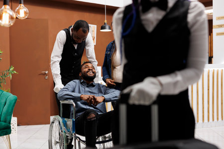 Wheelchair-friendly travel. Helpful hotel staff helping happy young male disabled guest at reception area, bellman pushing assisting man on wheelchair. Tourism and people with disabilitiesの写真素材