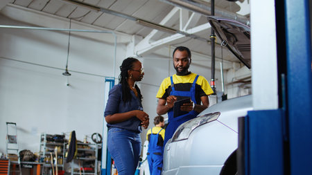 Trained repairman listens to customer in repair shop, using laptop to write down car modifications needed. Expert in garage with client, listening to her requests for vehicle tuning, ground level shotの写真素材