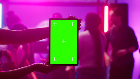 Woman holding greenscreen tablet at club and showing isolated display during dance party. Young funky adult using chroma key mockup template with blank copyspace on device screen.の写真素材