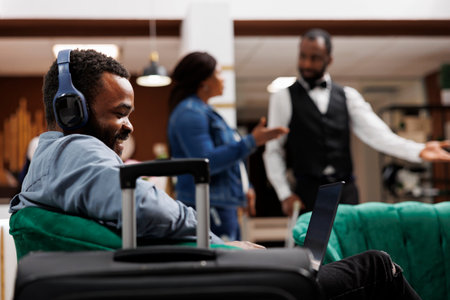 Happy African American man traveler wearing headphones looking at laptop screen. Hotel guest talking to family or friends on video call while traveling, sitting in lobby using computerの写真素材