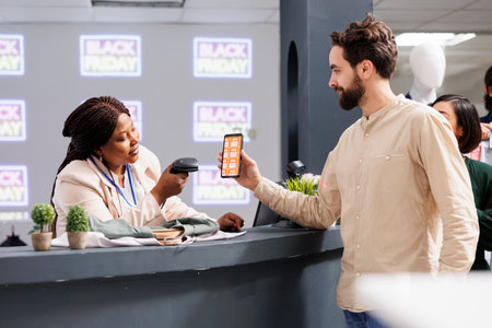 Happy young man standing at checkout desk in clothing store using mobile promo code to get Black...