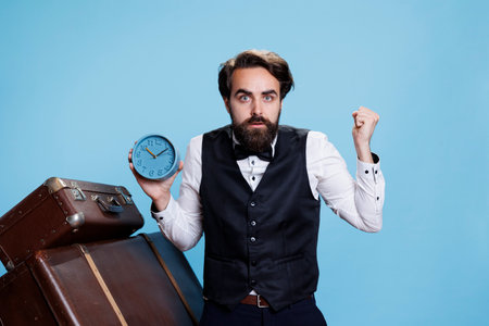 Hotel employee shows wall clock on camera, checking hour and overreacting for being late on duty. Professional doorman bellhop being punctual and looking at time on watch, blue background.の写真素材