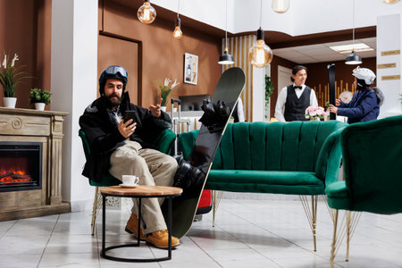 Youthful caucasian man in cozy hotel lobby during winter holiday using digital phone. Tourist wearing winter jacket and helmet sitting on sofa with snowboard and talking on mobile device.の写真素材
