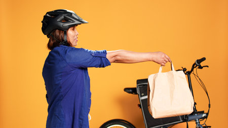 BIPOC food delivery worker reaching order address, waiting for customer to answer door. Courier woman greeting client, offering takeaway lunch bag, isolated over orange studio backgroundの写真素材