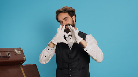 Cheerful bellhop does heart shaped sign in studio, expressing romantic honest feelings and flirting while he wears formal suit. Lovely employee showing romance symbol for love, sincere emotions.の写真素材