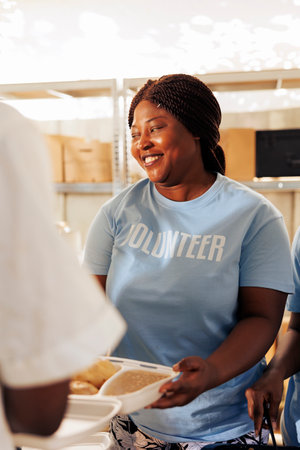 Friendly black woman giving free, hot meals to homeless people at a food drive. Portrait shot of african american female volunteer generously handing out food to the hungry and poor at a shelter.の写真素材