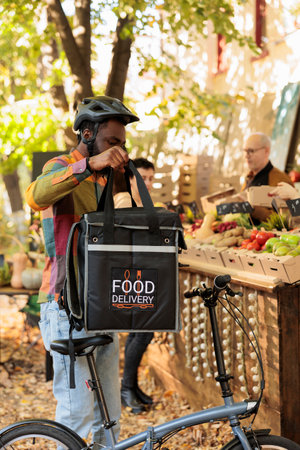 Young African delivery man standing near farm market stand picking up box with fresh organic produce. Black guy courier delivering fruits and vegetables produced by local farmers to customersの写真素材