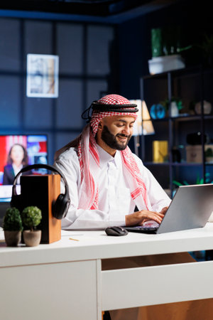 Arab businessman works diligently at his desk, typing on his digital laptop. Smiling Muslim guy using wireless technology for research and communication, showcasing his efficiency and professionalism.の写真素材