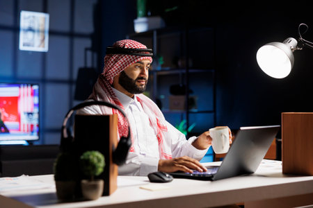 Side-view of a Muslim guy holding a cup of coffee and surfing the net on his digital laptop. Image...