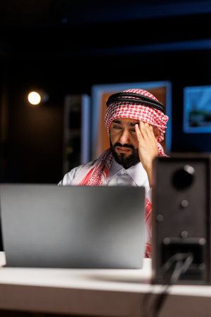 Detailed view of tired man in Arabian attire sitting at office desk and using laptop to conduct internet research. Muslim man with minicomputer on the table, looking weary and suffering from headache.の写真素材