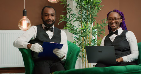 Hotel staff members reviewing guests list in lobby, reading information on forms and laptop. Team of employees managing reservations at luxury resort, tourism industry workers.の写真素材
