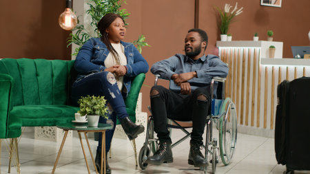 Wheelchair user hotel guest in lobby, african american man suffering from physical disability and going on vacation. Young couple at reception travelling despite reduced mobility. Handheld shot.の写真素材
