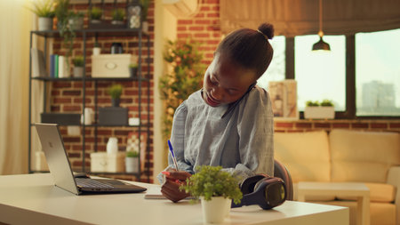 Woman takes handwritten notes while answering phone call at desk, writing important task information before starting job. Freelancer working through telephone chat and online apps.の写真素材