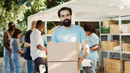 Generous man volunteering at neighborhood food drive, distributing non-perishable rations to the hungry and homeless. While holding donation box, caucasian charity worker looks at camera. Tripod shot.の写真素材