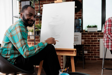 Art activities and wellbeing. Portrait of happy smiling African American guy during drawing lesson, young black man sitting at easel looking at camera, feeling inspired during sketching classの写真素材