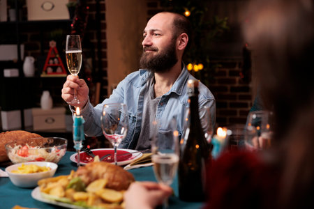 Young man raising glass for toast during christmas dinner at home, gathering with friends and family to eat traditional food and give speech at table. Joyful person enjoying festive december celebration.の写真素材