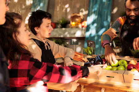 Young vendor helping client to choose fruits and vegetables, selling fresh organic homegrown produce at local farmers market. Woman talking to seller and buying natural seasonal food.の写真素材