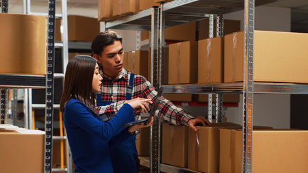 Team of workers analyzing cardboard boxes on shelves, checking merchandise stock in warehouse. Man and woman using laptop to work on products shipment, small business development.の写真素材