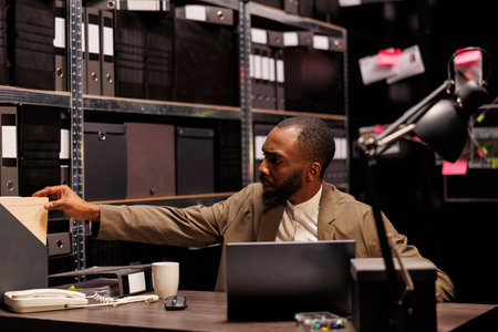 Detective analyzing crime case and searching police report while conducting investigation. African american policeman working in office and studying archival records in eveningの写真素材