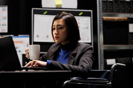 Paraplegic accountant businesswoman manager in wheelchair enjoying mug of coffee starting her shift in business administration workplace. Asian executive with disability sitting at office deskの写真素材